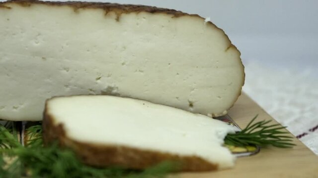 Round soft brown smoked homemade sliced cheese. Adyghe cheese with greens lying on wooden board . 4K