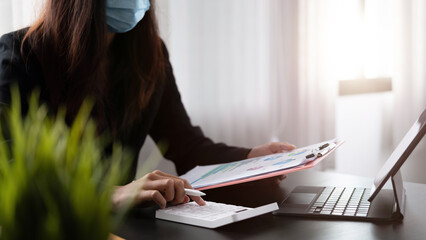 businesswoman wearing protective mask using calculator and calculating financial report in office