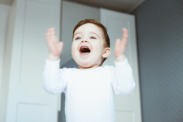 A portrait of a cute toddler baby boy in a white bodyuit seating on bed and laughing.Happy baby claps his hands, in bedroom at home