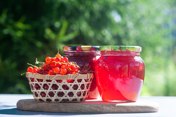 Red, juicy berries of red currants and jars of berry jam on a wooden table, on a green background of the garden, harvest and cooking theme