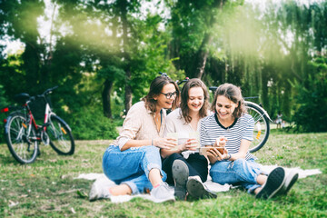Three young beautiful women friends having fun in a summer park sitting on green grass and looking into a mobile phone