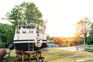 An old motor boat or yacht with two lifebuoys is moored in an outdoor workshop under repair next to dense green trees on a sunny summer day at sunset. Travel concept