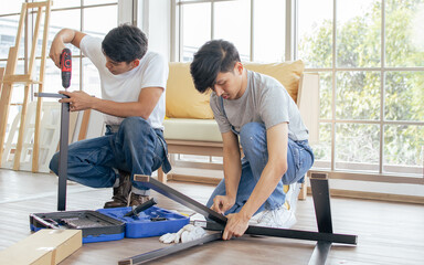 Two Asian handsome men wearing blue overalls, helping or supporting each other to assemble make DIY furniture following manual at home.