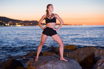 Glad woman is practicing stretching on a rock near sea at the dawn.