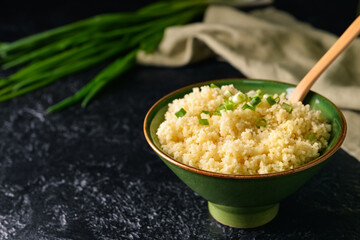 Bowl with tasty couscous and green onion on dark background