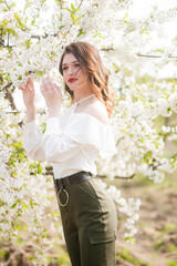 Fototapeta premium Lovely young girl in a white romantic silk blouse in spring in a blooming cherry garden in sunny weather. Spring and Fashion. Spring blooming gardens