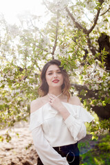 Lovely young girl in a white romantic silk blouse in spring in a blooming cherry garden in sunny weather. Spring and Fashion. Spring blooming gardens