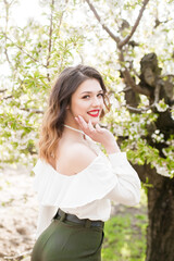 Fototapeta premium Lovely young girl in a white romantic silk blouse in spring in a blooming cherry garden in sunny weather. Spring and Fashion. Spring blooming gardens