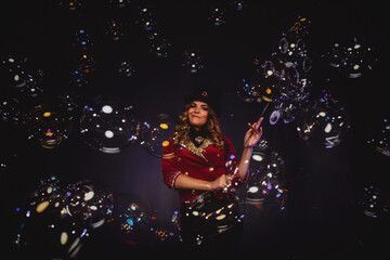 Female magician makes show with soap bubbles, an illusionist in theatrical clothes, on black background. Woman actress in stage costume and top hat on her head. Concept of performance. Copy space