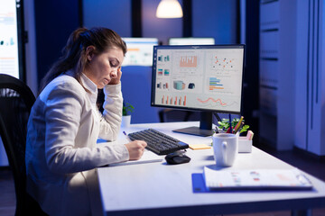 Back shot of overwhelmed woman working at night in front of computer, writing notes on notebook annual reports, checking financial deadline. Focused manager using technology network wireless