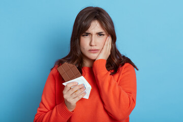 Upset Caucasian woman holding bar of chocolate in hands, feels toothache after biting sweets, covering cheek with palm, wearing casual sweater, isolated over blue background. Health care.