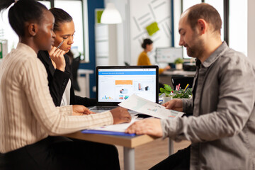 Busy multicultural, diverse employees analysing annual financial statistics sitting at desk in front of laptop holding documents searching business solutions. Team of businesspeople working in company