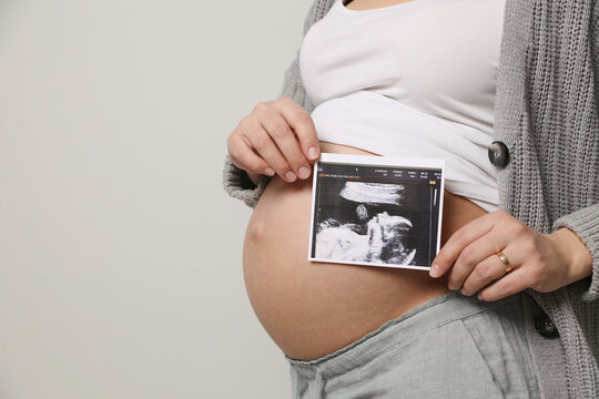 Young pregnant woman with ultrasound picture of baby on grey background, closeup. Space for text