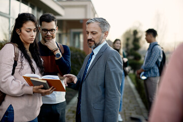 Mature teacher analysing lecture from a book with his students in front of the university.