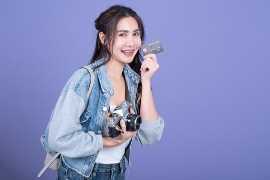 Cheerful Asian woman holding credit card ready to travel, tourism and vacation with backpack, photo camera on pink background.