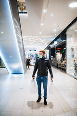 Full length of handsome caucasian man in black leather jacket with bags in hand standing in mall during epidemic COVID - 19 coronavirus