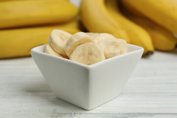 Bowl with cut bananas near whole fruits on white wooden table, closeup
