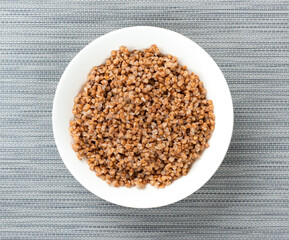 Freshly cooked buckwheat porridge in white plate on textured surface. Top view, closeup.