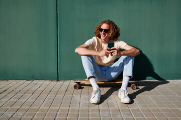 a young man using his smartphone and seated on his skateboard or longboard with a wall behind him