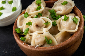 Meat dumplings in wooden bowl with sour cream and green onion on black background.