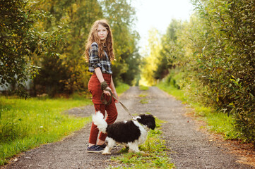 Naklejka na ściany i meble happy kid girl walking with her cavalier king charles spaniel dog on summer country road. Training her puppy and having fun.