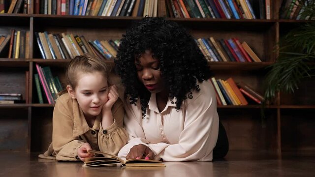 Afro-american stepmother and blonde adopted daughter read fairy tale in book smiling while sitting against large wooden bookcase at home