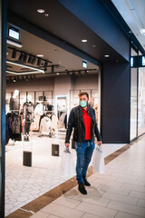 A young fashionable caucasian man in a leather jacket, with a mask and bags in his hand coming out of a boutique in a mall during an epidemic of the COVID-19 coronavirus 