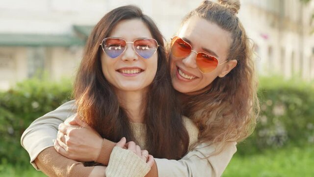 Portrait of two young smiling hipster women friends wearing sunglasses of heart shape. pretty girls hug, smile and show positive face emotions.