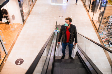 A young caucasian man with bags in his hand and a face mask on the escalator in the shopping mall. Shopping during the COVID pandemic - 19 coronavirus