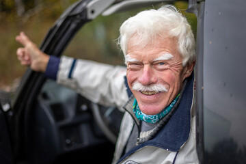 Close-up portrait of a happy, laughing older man in his car standing on the forest path.