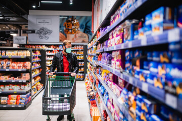 A young man with a medical face mask pushes a basket to the supermarket and looks at groceries. Procurement of food during the coronavirus pandemic COVID - 19