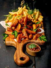 Appetizing fried potato wedges on a wooden cutting board. Big portion of baked potatoes with sauce and herbs. Homemade french fries organic potato slices. Fast food photography. Vertical shot