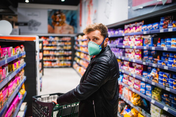 The back view of a Caucasian young handsome man with a face mask buys food at the supermarket and pushes a grocery basket. Procurement of food during the coronavirus pandemic COVID - 19