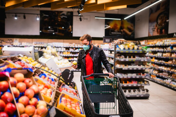 A Caucasian fit man with a face mask who buys healthy food at the supermarket and pushes a grocery basket. Procurement of food during the coronavirus pandemic COVID - 19
