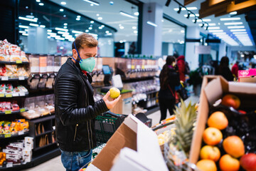 A Caucasian man with a face mask holding an apple in his hand and buying healthy food at the supermarket. Procurement of food during the coronavirus crisis COVID - 19 pandemic