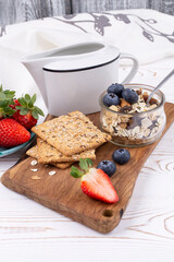 Muesli with nuts, fresh strawberries and blueberries, milk jug, crispbread on a white wooden table in a rustic style, close-up, vertically in light colors