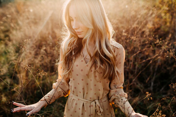 Young woman with long blonde wavy hair, at sunset, walking in a field with tall dry grass.