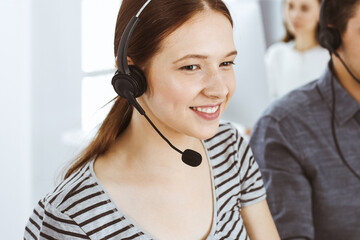 Casual dressed young woman using headset and computer while talking with customers online. Group of...