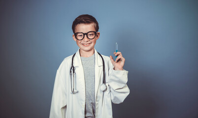 cool young boy is disguised as a doctor with a white doctor smock and plays with syringe and stethoscope in front of blue background in the studio