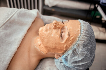 The client's face is a young woman with an alginic orange mask. Thick mask. Professional beauty salon.