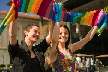 A young lesbian couple embraces their love. They are walking the streets and waving with a rainbow colors scarf.