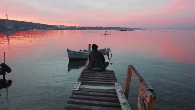 Lonely Refugee Man Sits on the Wooden Pier at Levos Island Port, Red Sky Sunset in Background