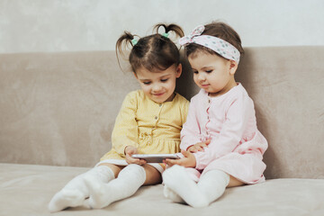 Happy little girls sisters sit on couch in the living room and watch cartoons on smartphone together.