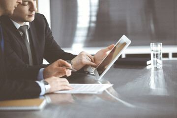 Unknown businessmen using tablet computer and work together at the glass desk in modern office, close-up. Teamwork and partnership concept