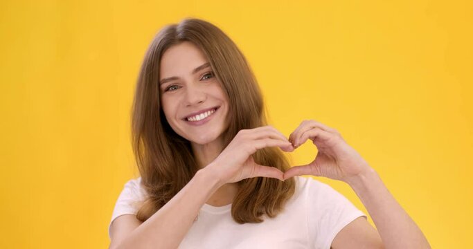 Kindness concept. Young cute positive lady showing heart shape with her hands and smiling to camera, orange background