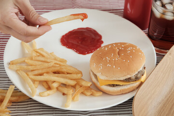 Close-up shot, selective focus: Man's hand was eating French fried, dipping ketchup on a white plate on red tablecloth with hamburgers and cola. Eating junk food or fast food for lunch is unhealthy.