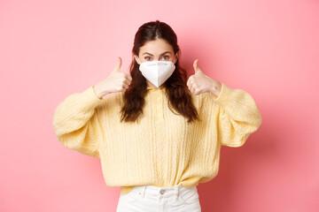 Covid-19, lockdown and pandemic concept. Young woman wearing respirator, face mask during quarantine, showing thumbs up in approval, support vaccination, pink background - obrazy, fototapety, plakaty