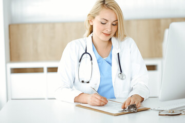 Woman-doctor at work while sitting at the desk in hospital or clinic. Blonde cheerful physician filling up medication history record form
