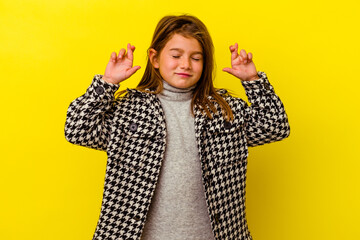 Little caucasian girl isolated on yellow background crossing fingers for having luck