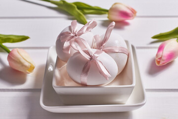 Three white eggs with pink bows on a porcelain plate on a white wooden background and tulip flowers.